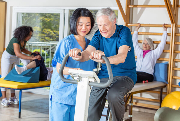 Asian nurse helping elder man in hospital gym.