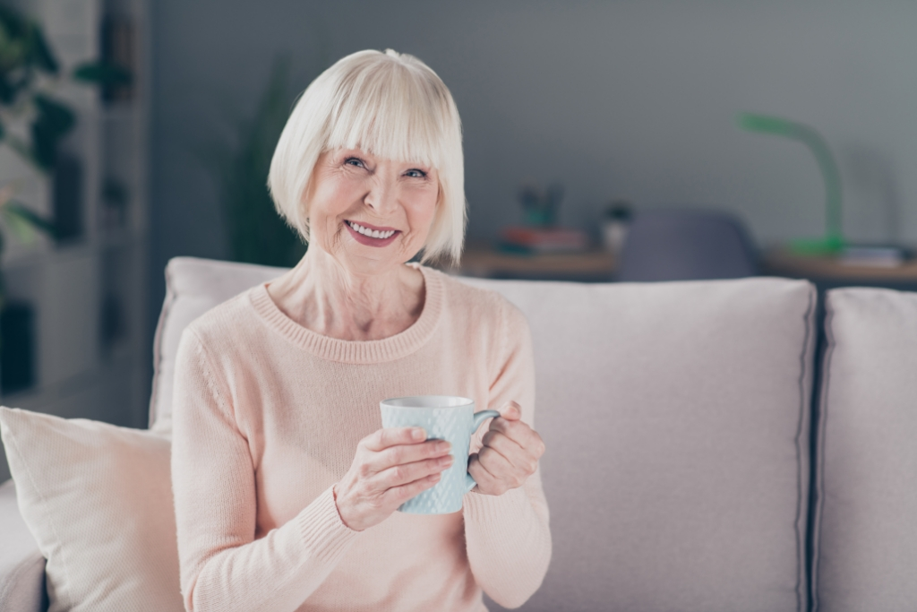 senior woman holding a cup of coffee, sitting on the sofa in her assisted living apartment, before she starts a full day of senior living activities