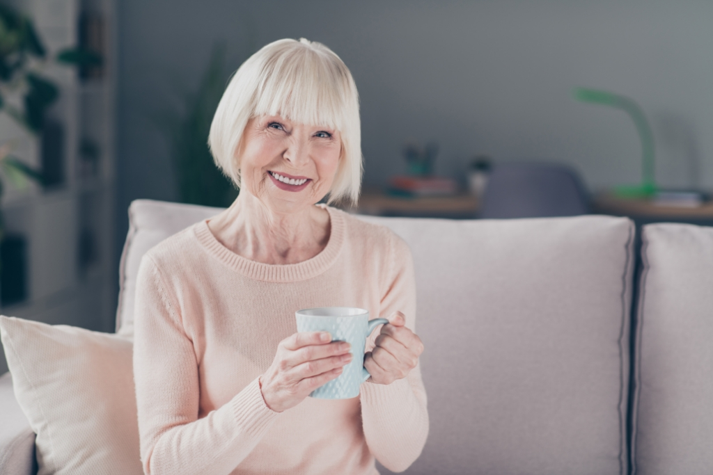 Senior woman holding a cup of coffee, sitting on the sofa in her assisted living apartment.